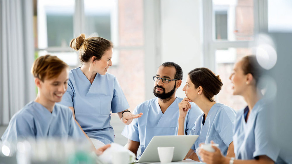 Medizinisches Team in hellblauen OP-Kleidern sitzt um einen Tisch in einer hellen Krankenhausumgebung und bespricht die Patientenversorgung mit einem Laptop und Kaffeetassen auf dem Tisch