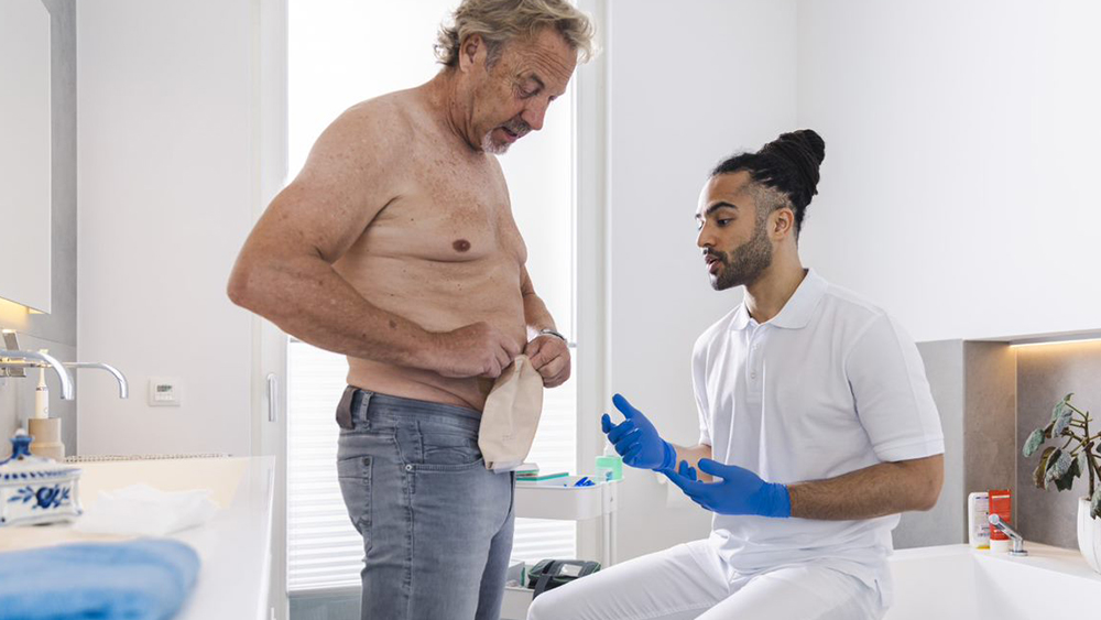 Mann mit nacktem Oberkörper im privaten Badezimmer manipuliert einen Stomabeutel auf dem Bauch. Eine männliche Pflegekraft sitzt auf dem Badewannenrand und gibt Anweisungen.