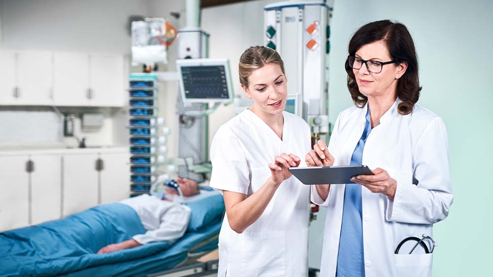 Un médecin et une infirmière discutent de la posologie de la perfusion intelligente B. Braun, qui est saisie directement à partir d’une tablette. Le patient en unité de soins intensifs peut être vu en arrière-plan, connecté à une tour remplie de dispositifs de perfusion intelligents.