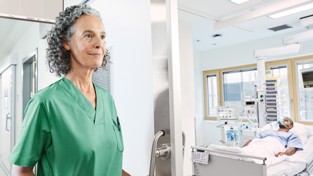 Une personne portant des pyjamas médicaux se tient à l’extérieur d’une salle de soins intensifs avec un patient dans un lit d’hôpital.