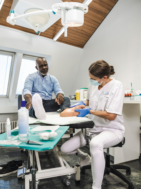 Un professionnel de santé portant des gants traite le pied d’un patient sur une table d’examen dans une salle de clinique, avec des fournitures médicales sur un plateau à proximité et une lampe d’examen suspendue au-dessus.