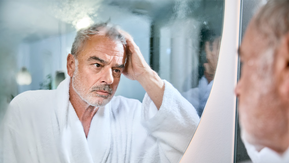 un homme âgé en peignoir regarde attentivement dans le miroir de la salle de bain embué